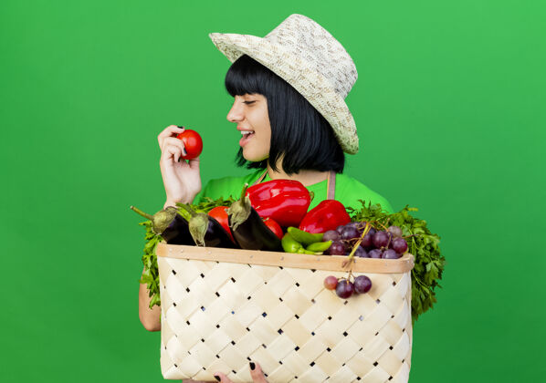 制服穿着制服 戴着园艺帽的快乐的年轻女园丁拿着菜篮子 看着西红柿花园帽子蔬菜