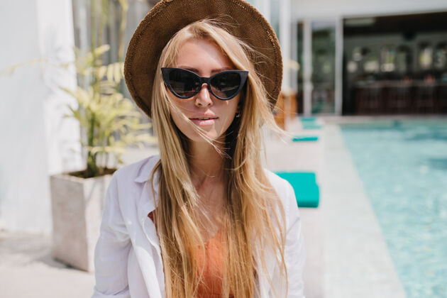 金发漂亮的白人模特戴着棕色帽子坐在度假村的游泳池旁严肃的美女戴着黑色太阳镜 享受着夏日的好天气情感阳光年轻