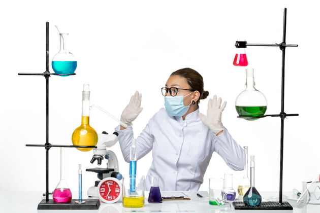 专业前视图穿着医疗服的女化学家带着面罩坐在桌子前 白色背景上有溶液病毒化学实验室的covid飞溅物坐药人