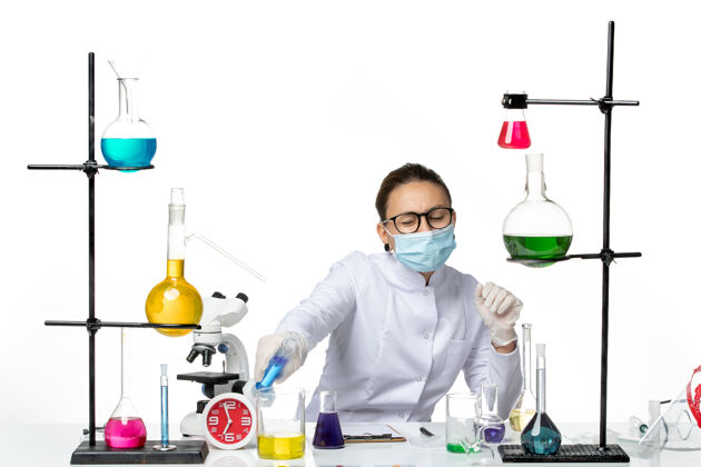 前面前视图穿着医疗服的女化学家戴着口罩坐在房间里 桌子上放着白色背景上的溶液病毒化学实验室covidsplash解决方案里面病毒