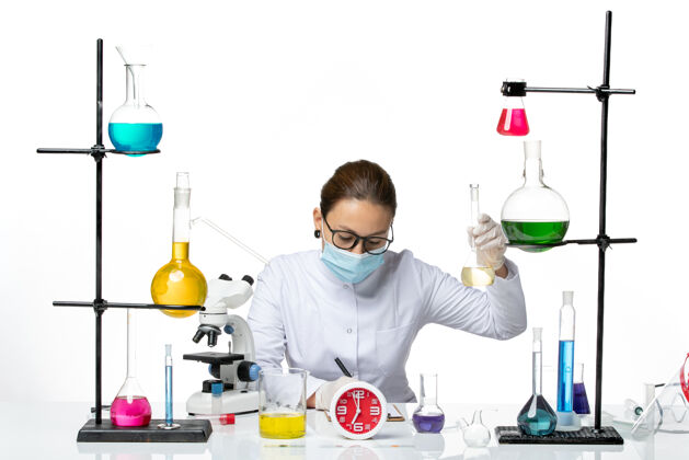 喷溅前视图穿着白色医疗服的女化学家 在桌子前面戴着面罩 溶液背景为浅白色化学家实验室病毒冠状病毒飞溅套装面罩医生