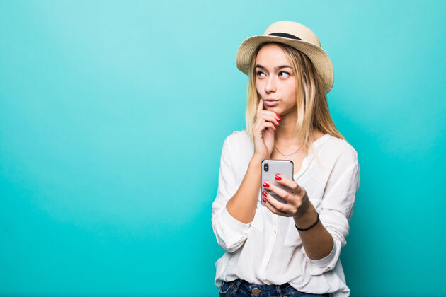 深褐色想着戴草帽的年轻女子用手机隔着蓝色的墙华丽滚动互联网