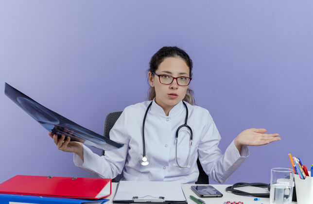 女性困惑的年轻女医生穿着医用长袍 听诊器坐在办公桌旁 手持医疗工具 拿着x光片 看样子空手孤立桌子表演长袍