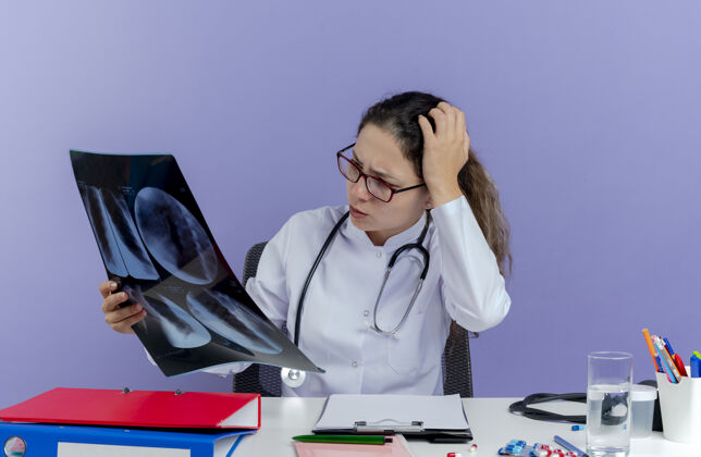 射击困惑的年轻女医生穿着医用长袍和听诊器坐在办公桌旁 手里拿着医疗工具 看着x光片 手放在头上 与世隔绝女性保持长袍