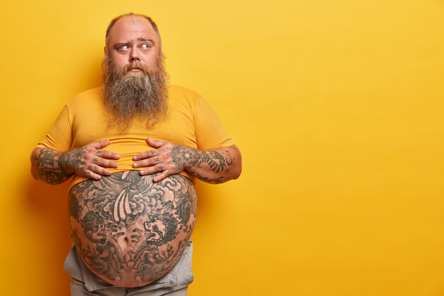 空超重沉思的男人双手放在大肚子上纹身 一边思考 一边看 留着浓密的胡须 靠着黄色的墙壁摆姿势肥胖的男人不知道肚子怎么会出现肥胖空白大