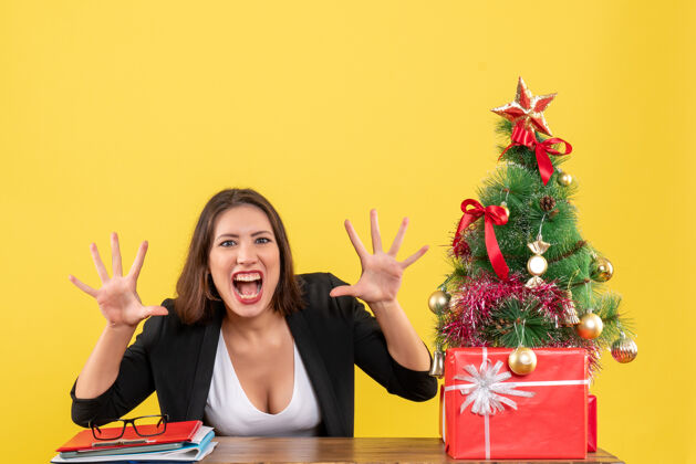 女人震惊的年轻女子显示十个坐在一张桌子旁装饰圣诞树在黄色办公室桌子年轻女人漂亮