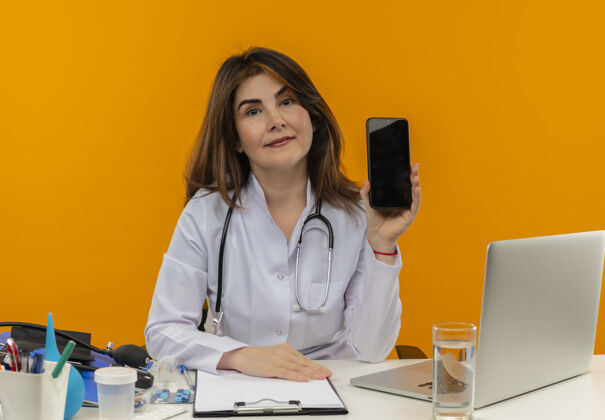 橙色高兴的中年女医生穿着医用长袍和听诊器坐在办公桌前 用医疗工具剪贴板和笔记本电脑显示手机被隔离电话手机长袍