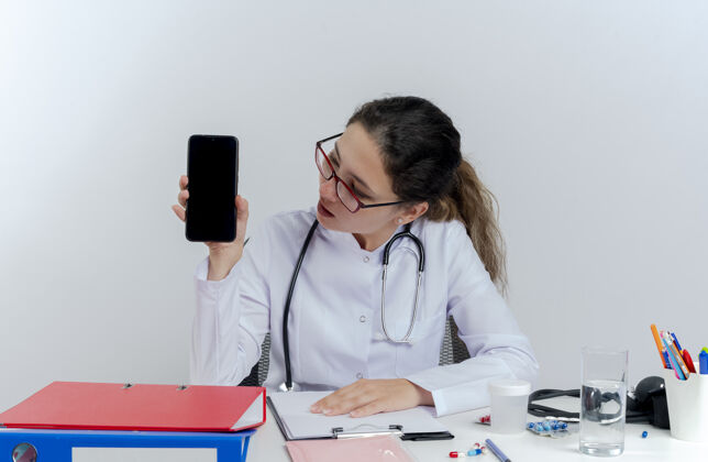 成人令人印象深刻的年轻女医生穿着医用长袍和听诊器 戴着眼镜坐在办公桌前 拿着医疗工具展示和看手机年轻女性工具