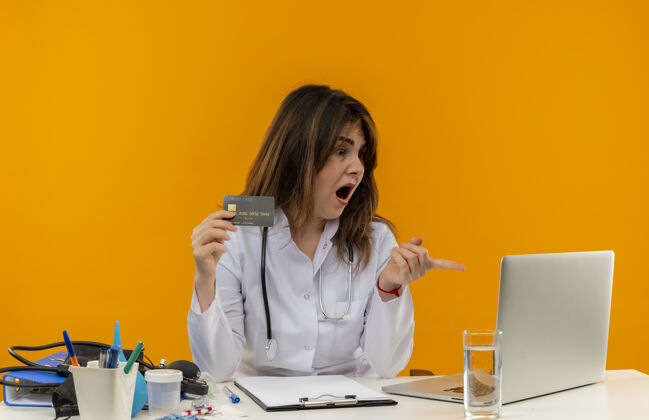 惊喜惊讶的中年女医生穿着医用长袍和听诊器坐在办公桌旁 拿着医疗工具剪贴板 看着并指着手提电脑 手里拿着信用卡笔记本电脑指着办公桌