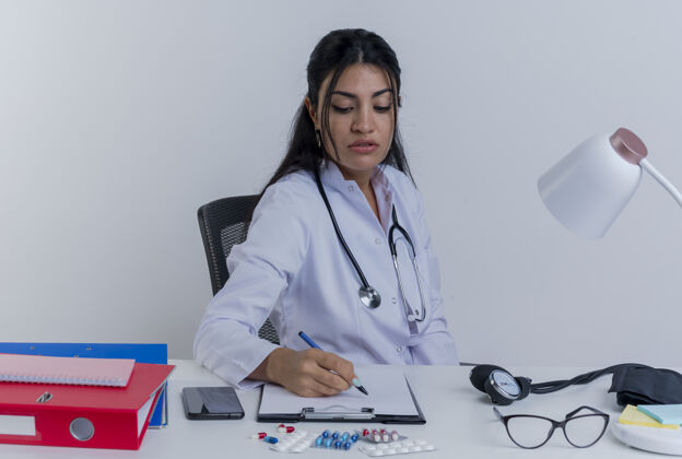 专心专注的年轻女医生穿着医用长袍 听诊器坐在办公桌旁 医疗工具写在剪贴板上女人桌子长袍