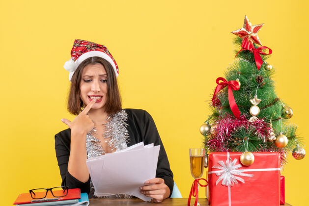 大人困惑的商务女士穿着西装 戴着圣诞老人的帽子和新年装饰品 手里拿着文件 坐在一张桌子旁 桌子上放着圣诞树年抱圣诞老人