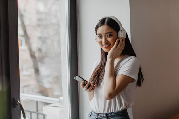美丽一位身穿白色上衣的棕色眼睛的亚洲女性微笑着看着前方 手持智能手机 戴上耳机休闲魅力科技