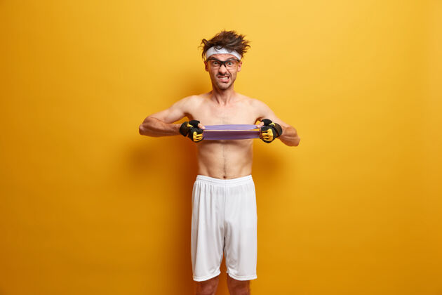 肌肉运动强人拉弹力阻力带 锻炼手部肌肉 进行健身健美训练 戴运动手套和白色短裤 隔离在黄色墙壁上健康的生活方式力量肌肉年轻