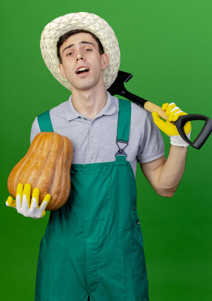手套自信的年轻男性园丁戴着园艺帽和手套 肩上拿着南瓜和铁锹 在绿色背景上与复制空间隔离开来肩膀男年轻