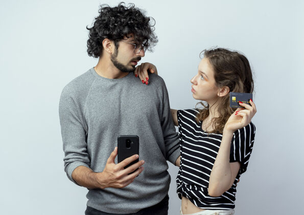 信用年轻漂亮的情侣男女 迷茫的男人拿着智能手机 看着女朋友拿着他的信用卡 背景是白色的流动夫妻持有