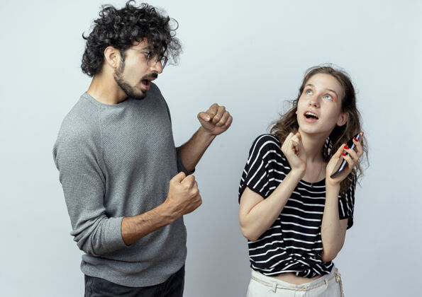 女孩一对年轻的男女 失望的男人紧握拳头看着拿着智能手机站在白色背景下的惊讶女孩拳头年轻移动