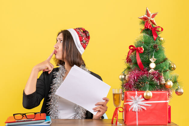 抱着一个穿着西装 戴着圣诞老人帽 戴着新年装饰品 一个人拿着文件 坐在办公桌旁 桌上放着圣诞树的困惑的商务女士圣诞老人帽子时尚