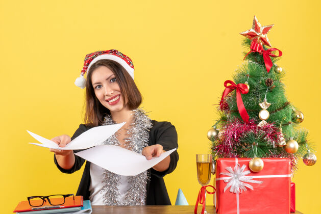 坐着穿着西装 戴着圣诞老人帽和新年装饰品 展示文件的商务女士 坐在办公室的桌子旁 桌上放着圣诞树成人圣诞老人桌子