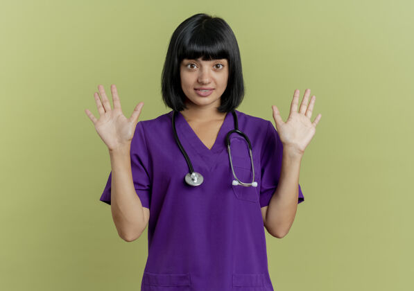 空间惊讶的年轻黑发女医生穿着制服 手持听诊器站在橄榄绿的背景上 双手分开 留有复印空间年轻站复制