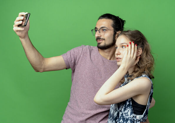 拍摄一对年轻的男女 快乐的男人用他的智能手机站在绿色的背景上给他们拍照站立照片手机