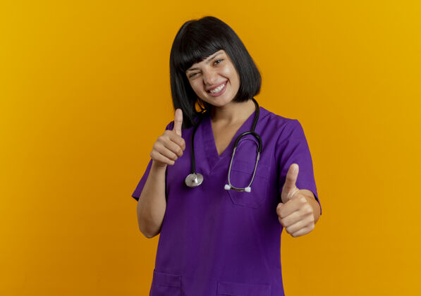 橙色微笑着的年轻黑发女医生穿着制服 手持听诊器 竖起大拇指 两手分开放在橙色背景上 留有复印空间年轻听诊器拇指