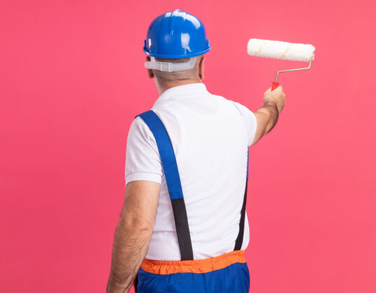 抱自信的成年建筑工人穿着制服站着 从后到前拿着滚筒刷隔离在粉红色的墙上衣服建设者刷子