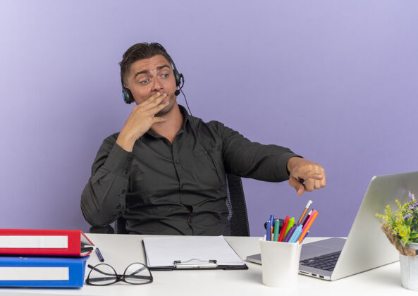 办公桌年轻惊讶的金发上班族戴着耳机坐在办公桌旁 拿着办公工具 看着笔记本电脑 把手放在嘴上 在紫色背景上 留着复印空间金发耳机笔记本电脑