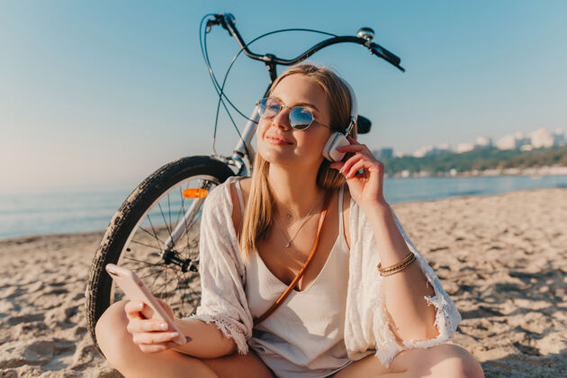 服装时尚年轻迷人的金发微笑的女人坐在沙滩上骑着自行车戴着耳机听音乐女性阳光音乐