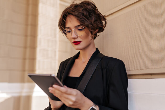 华丽黑发女人 红唇在外面摆姿势 手持平板电脑时髦女人 短发 黑色套装 戴眼镜 在户外摆姿势走道街道城市
