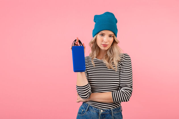 女人年轻漂亮的女人拿着无线扬声器听音乐穿着条纹衬衫和蓝色帽子微笑快乐积极的心情摆在粉红色的墙上服装乐趣享受