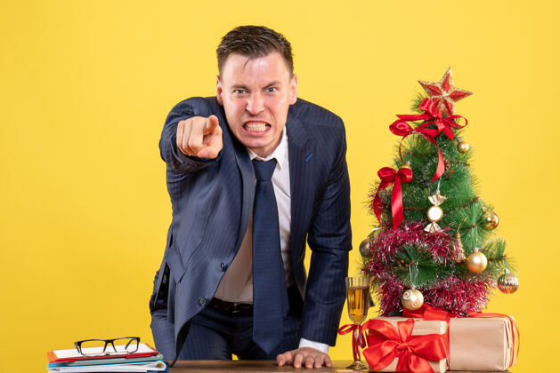 业务员前视图愤怒的商人站在圣诞树旁的桌子后面 在黄色背景上呈现圣诞节办公室人