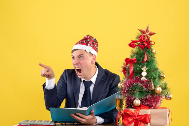 笔记本电脑前视图愤怒的商人对着坐在圣诞树旁桌子上的人大喊大叫 黄色背景的礼物行政业务员男