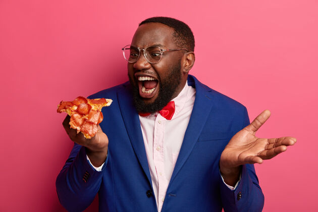 不健康饥肠辘辘的黑人咬着一大块比萨饼 胃口大开 穿着正装戴着眼镜对着粉色空间摆姿势正式的衣服垃圾嘴巴