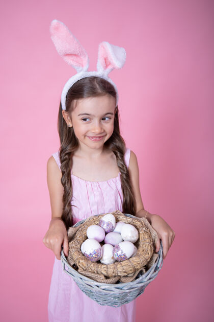 彩蛋小女孩带着复活节兔子耳朵 拿着一个篮子 篮子里放着复活节彩蛋 在粉色的墙上特写复活节彩蛋孩子节日