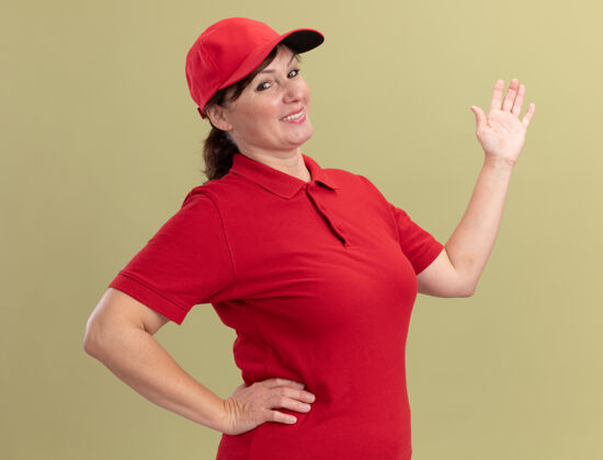 年龄身穿红色制服 头戴帽子的中年送货妇女站在绿色墙壁上 望着前方展示复印空间展示中间站立