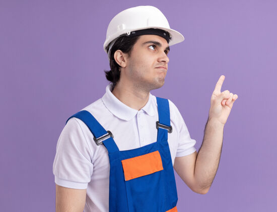 安全年轻的建筑工人穿着建筑制服 戴着安全帽 看上去很困惑 用食指指着站在紫色墙上的一边混乱人建筑