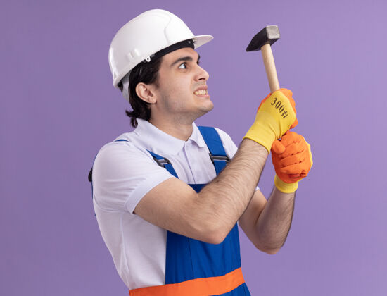 建筑愤怒的年轻建筑工人身穿建筑制服 戴着橡胶手套戴着安全帽 手持锤子 站在紫色的墙上 带着恼怒的表情看着它男人橡皮表情