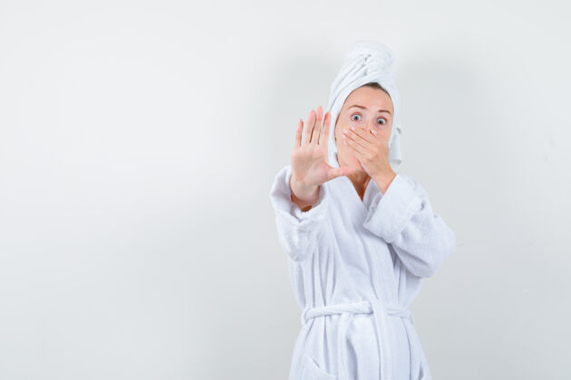 手势照片中的年轻女子手捂着嘴 穿着白色浴衣 毛巾做着停车的手势 看上去很害怕乐趣浴袍肖像