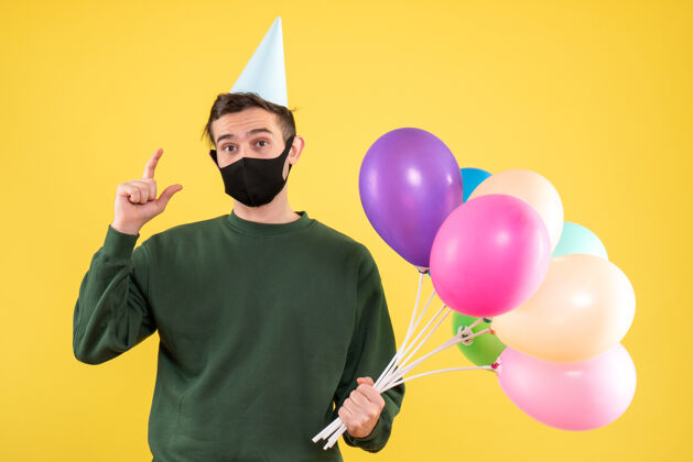 乐趣正面图：戴着派对帽的年轻人 戴着黄色背景的黑色面具和五颜六色的气球快乐背景年轻人