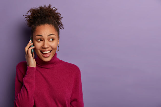 卷发高兴的黑人妇女有电话交谈 看一边 和密友讨论喜讯 打电话给某人 穿紫红色高领毛衣高兴手机满意