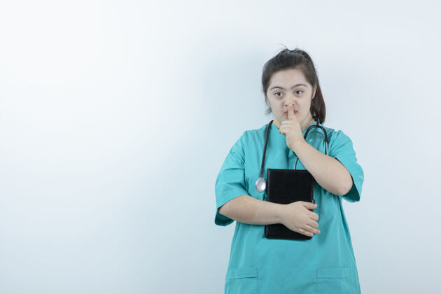 笔记本年轻的女护士 手持听诊器和记事本 在白色的墙上显示无声的手势安静医生女孩