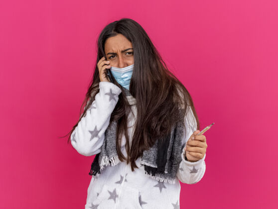 围巾未被释放的年轻生病的女孩看着相机戴着医用面罩和围巾讲电话 拿着隔离在粉红色背景上的温度计年轻电话穿着