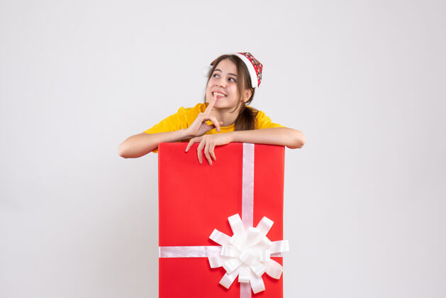 购物前视图可爱的女孩与圣诞老人帽子站在大礼物后面欢呼圣诞节人