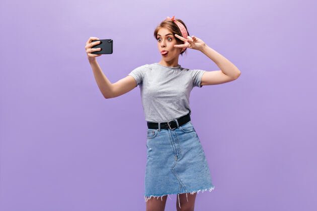 电话穿着牛仔裙的女孩展示舌头 展示和平标志 自拍风趣的女人穿着时髦的衣服摆姿势 心情很好脸紫色复制