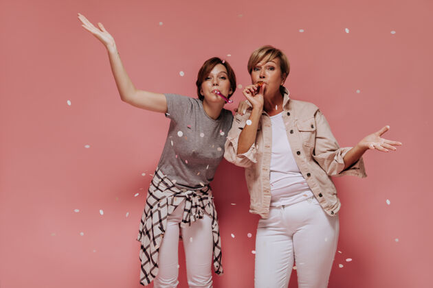 年轻时尚的两位女士 现代短发 穿着白色裤子 看着镜头 在粉色背景下摆出五彩纸屑的姿势礼物酷中等
