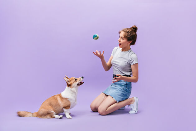 肖像女孩坐在地板上和狗玩耍穿着白色运动鞋的快乐的年轻女子在紫色背景上摆着球和高尔基犬的姿势头发女孩表情