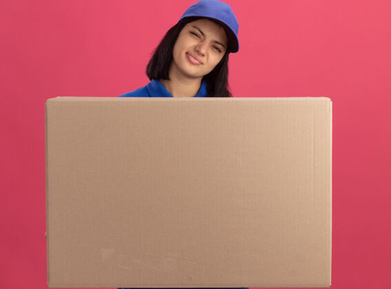 使身穿蓝色制服 戴着帽子的年轻送货员站在粉红色的墙上 手里拿着一个大纸箱 嘴角歪歪扭扭的 脸上露出失望的表情年轻表情制服