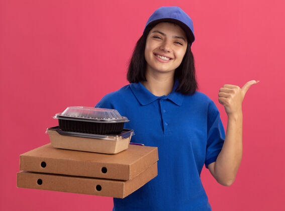 微笑身穿蓝色制服 头戴鸭舌帽 手持比萨饼盒和食品包的年轻送货女孩微笑着用拇指指着站在粉色墙上的一边盒子包装制服