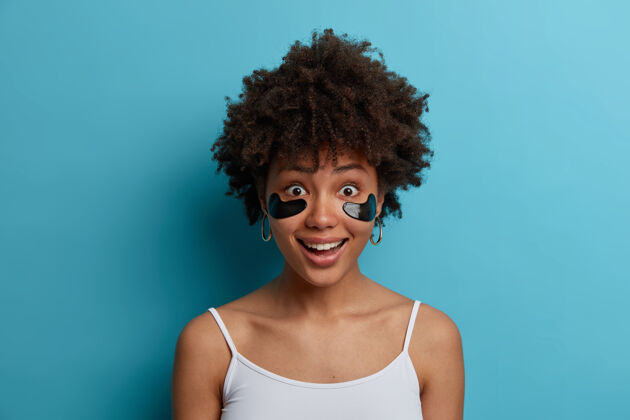 年轻快乐的黑皮肤女人的近景拍摄有抗衰老的眼部治疗 在眼部应用化妆品贴片 想要有健康的皮肤 穿便服 隔离在蓝色的墙壁上反应治疗背心