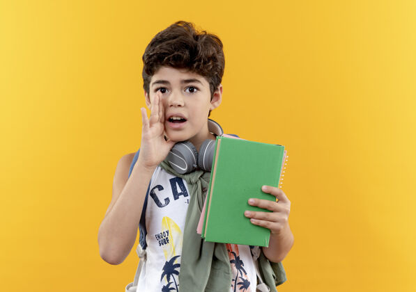 包看着摄像机 一个背着书包 戴着耳机 拿着书 在黄色背景上低语的小男孩戴男孩背